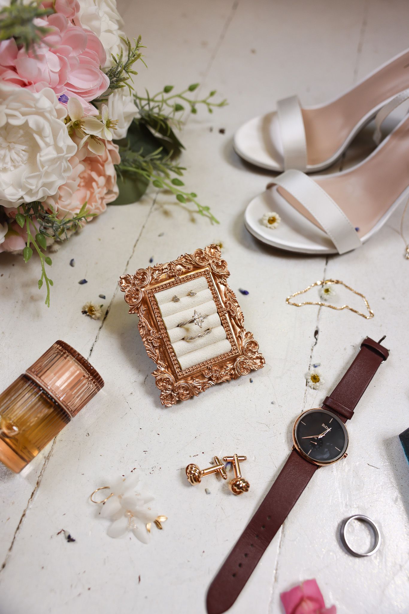 Wedding accessories including rose gold frames, watches, and floral arrangements on a white surface.