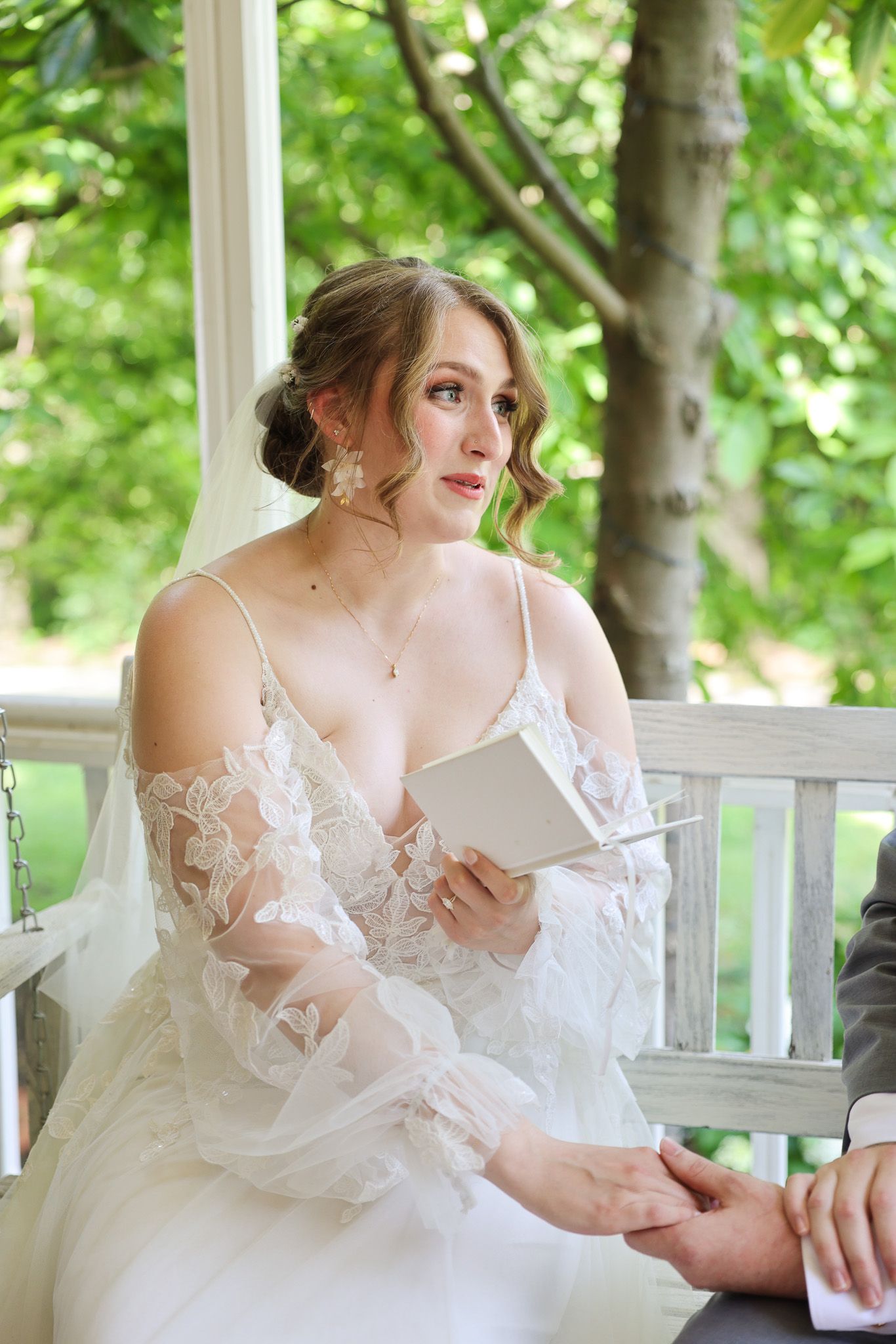 A person in a white lace wedding dress reads letters on a porch surrounded by greenery.