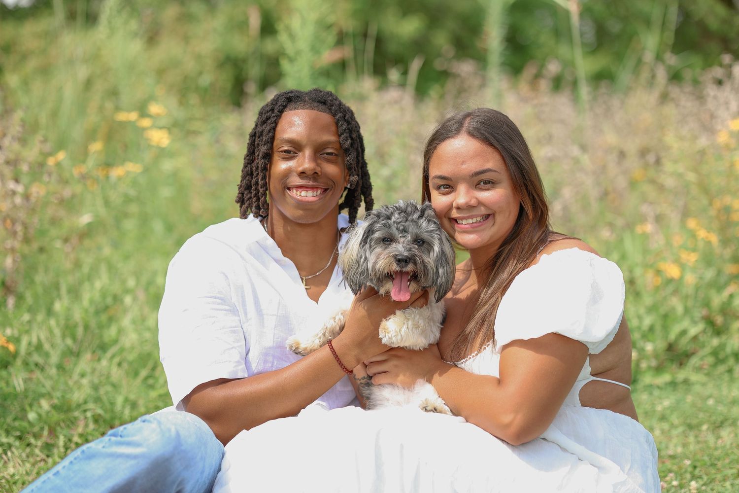 A couple in white outfits holds their small gray dog during an outdoor photoshoot.