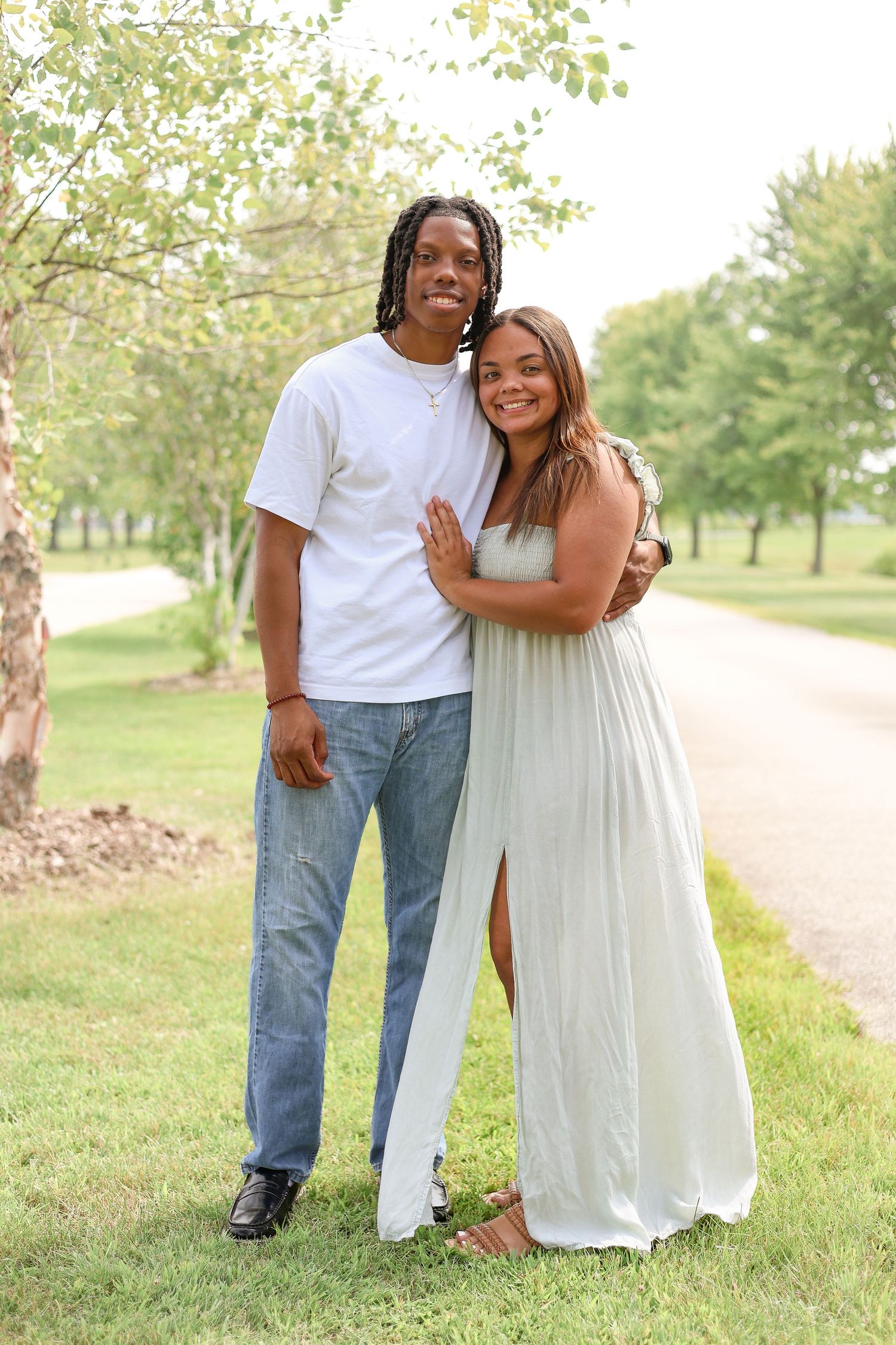 Happy couple embracing outdoors on a sunny day, wearing casual white attire on a tree-lined path.