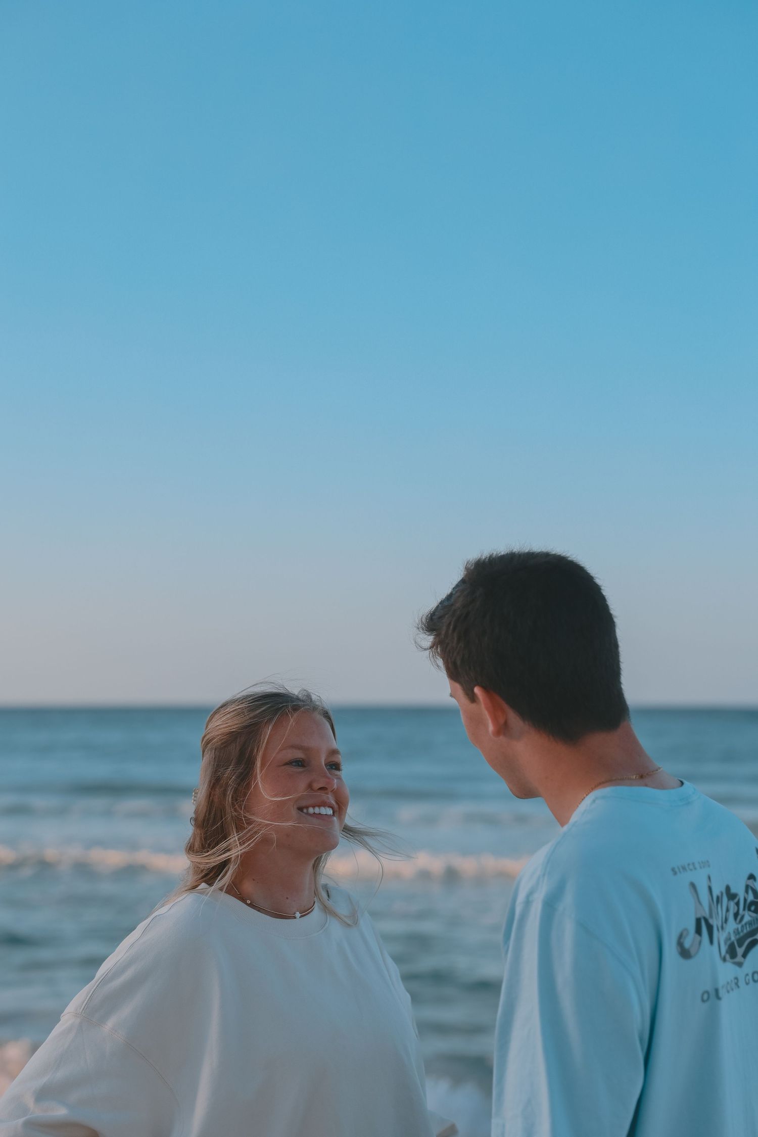 Smiling blonde woman and man sharing a moment on a beach at sunset.