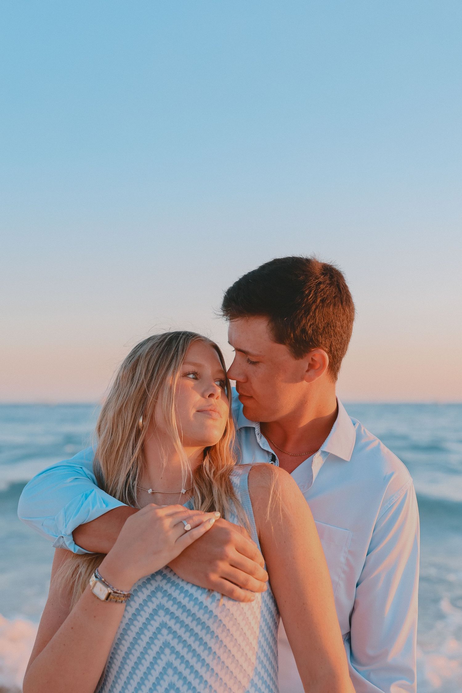 Young couple sharing a romantic moment at beach sunset, man embracing woman from behind while gazing into each others eyes.
