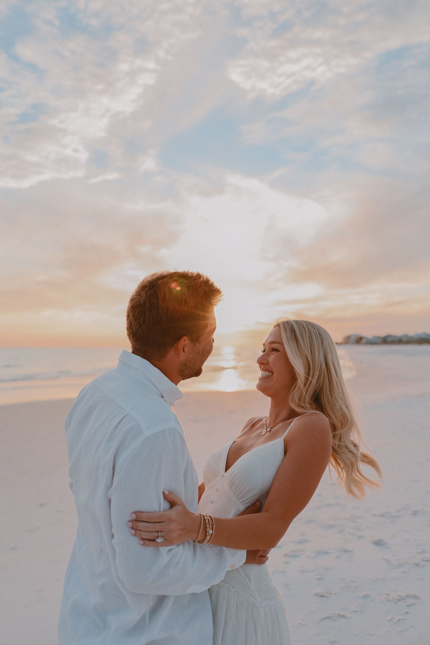 Happy engaged couple embracing on sandy beach at golden sunset, woman laughing joyfully in white dress.
