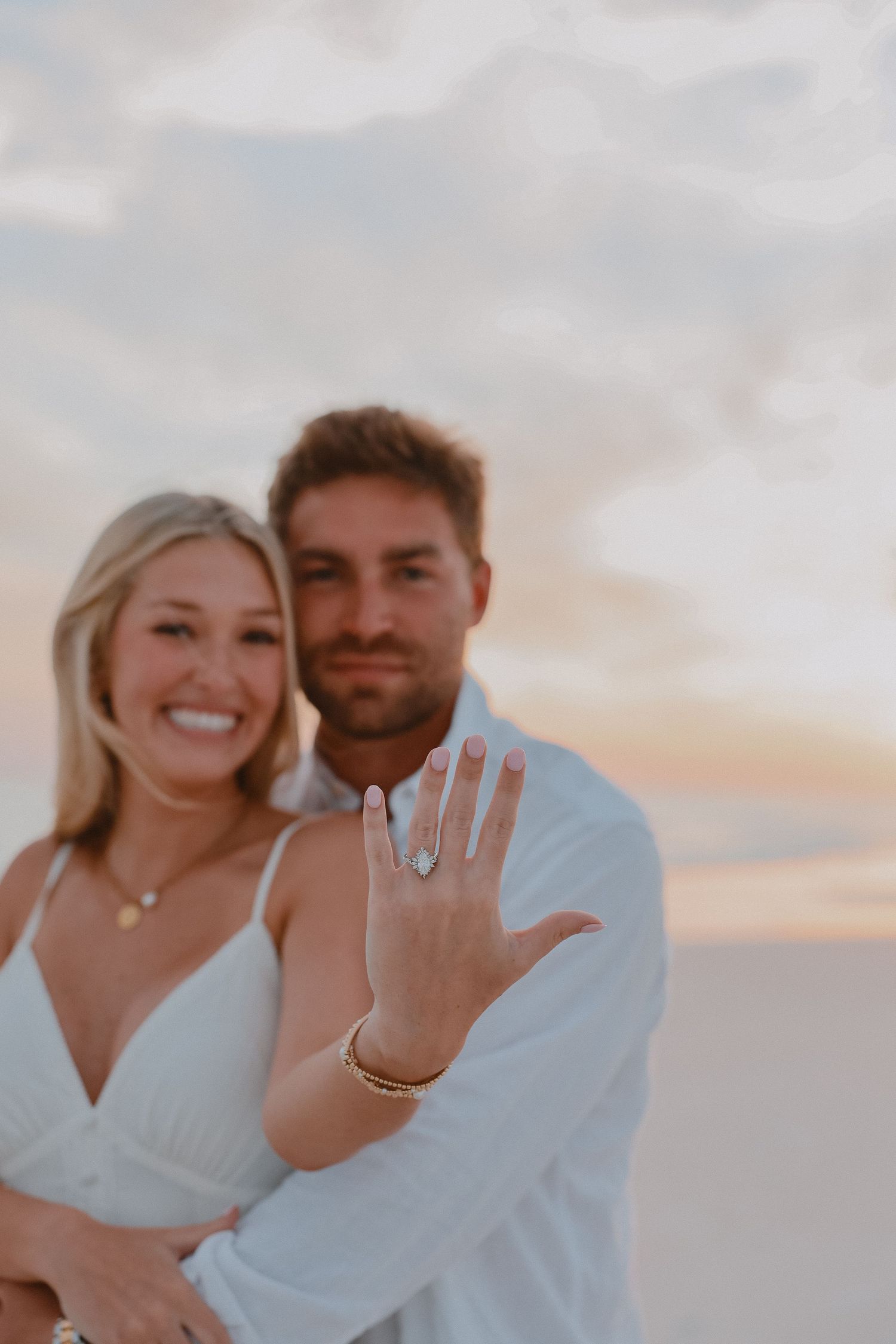 Smiling engaged couple showing off diamond engagement ring on beach at pastel sunset.