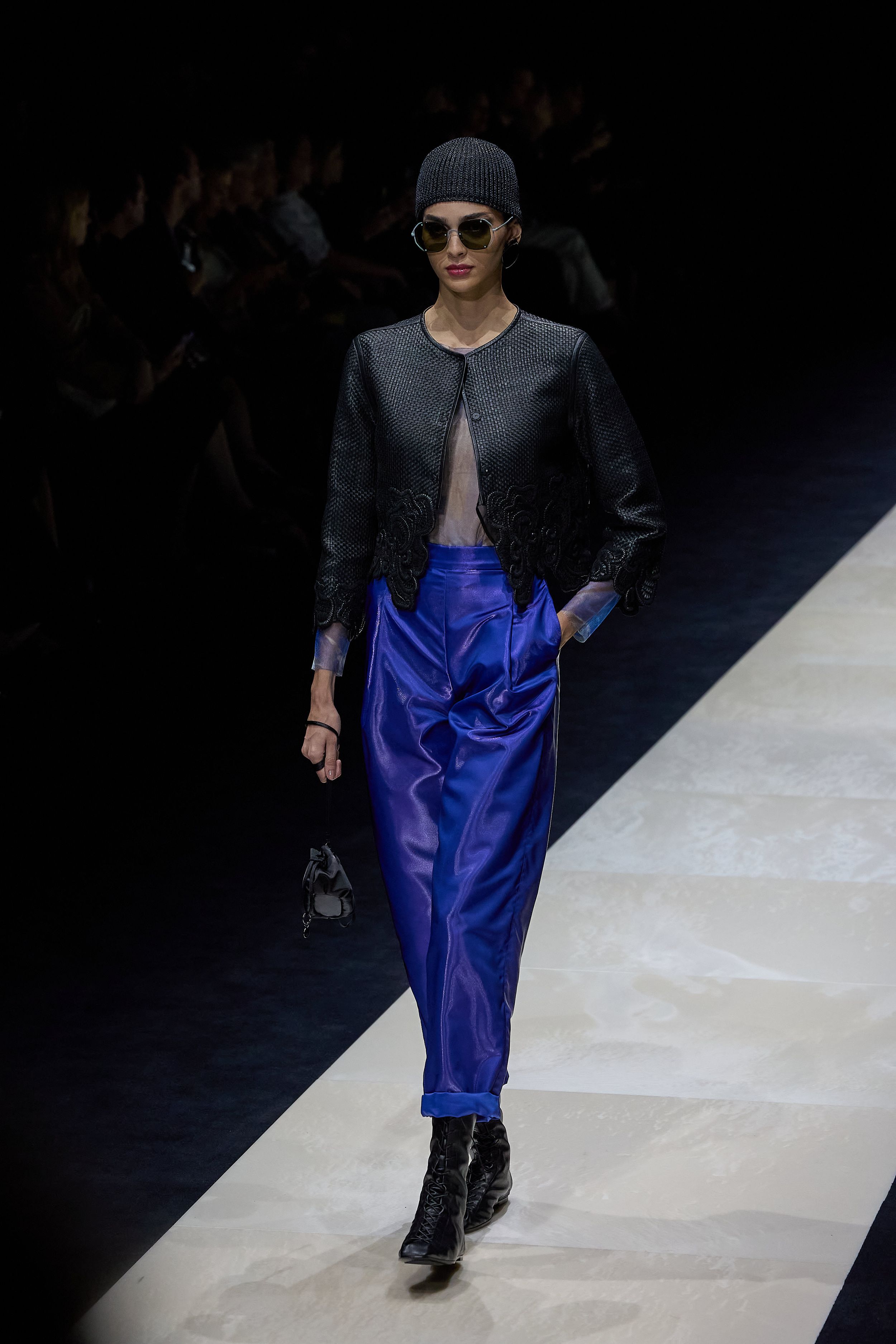 Fashion model walking runway in black leather jacket and royal blue pants against dark background.