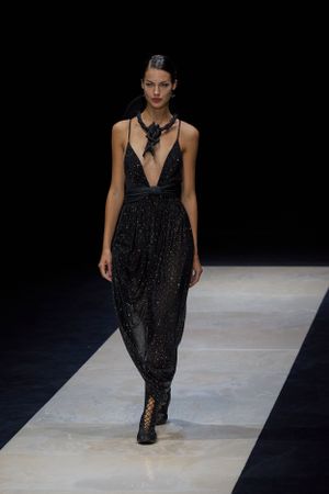 Sparkly black evening gown with plunging neckline and statement necklace on runway.