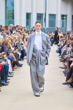 Oversized gray suit ensemble with white shirt shown on outdoor runway with seated audience.
