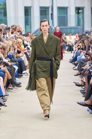 Olive military-style coat with fringe detail and khaki pants shown on runway with audience.