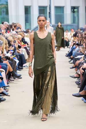 Model showcases olive tank dress with dramatic floor-length fringe at fashion week runway show.