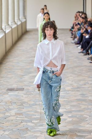 Runway model wearing cropped white shirt and embellished denim jeans with green platform shoes.