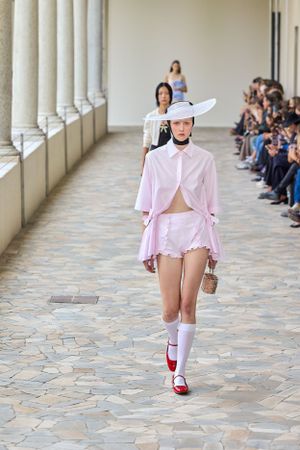 Fashion model walks runway in pink ensemble with white sun hat and knee-high socks during outdoor fashion show.