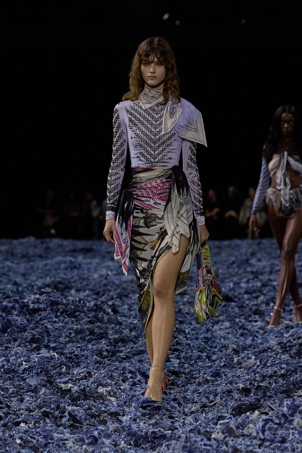 Model presents flowing dusty rose maxi dress with ruffled layers on dark runway.