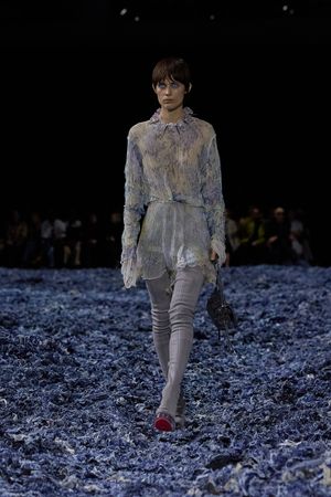 Fashion model walks runway in delicate lace dress with high collar over gray pants against dark backdrop.
