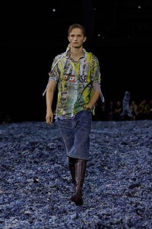 Model wearing patterned shirt and cropped jeans walks on blue textured runway surface.