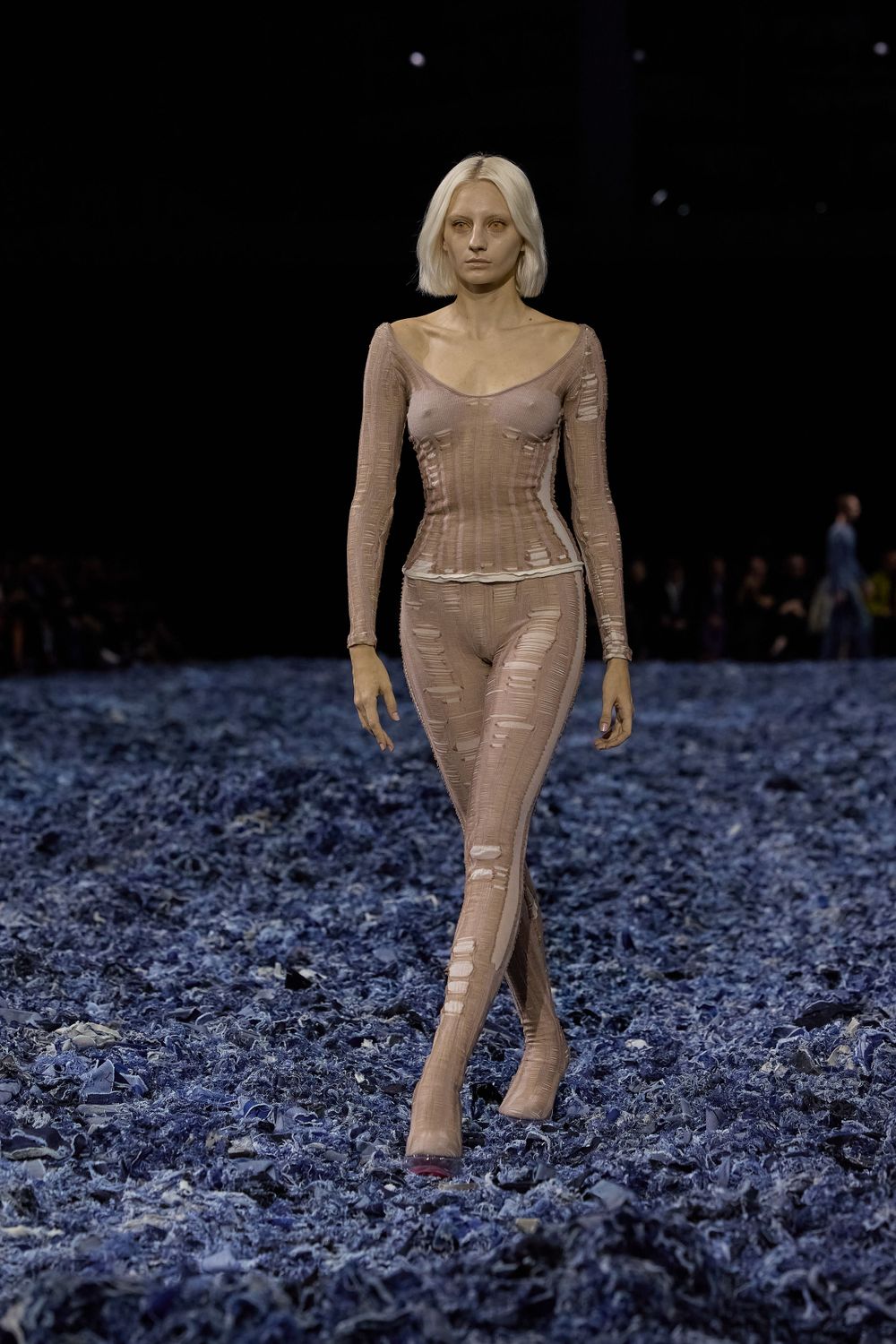 Model presents form-fitting nude bodysuit on dark runway with blue textured floor.