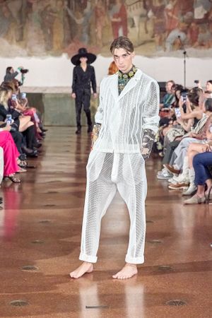 Model showcases white mesh ensemble with bare feet on runway with mural backdrop.