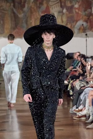 Model wearing black textured suit and wide-brimmed hat walks runway at fashion show.
