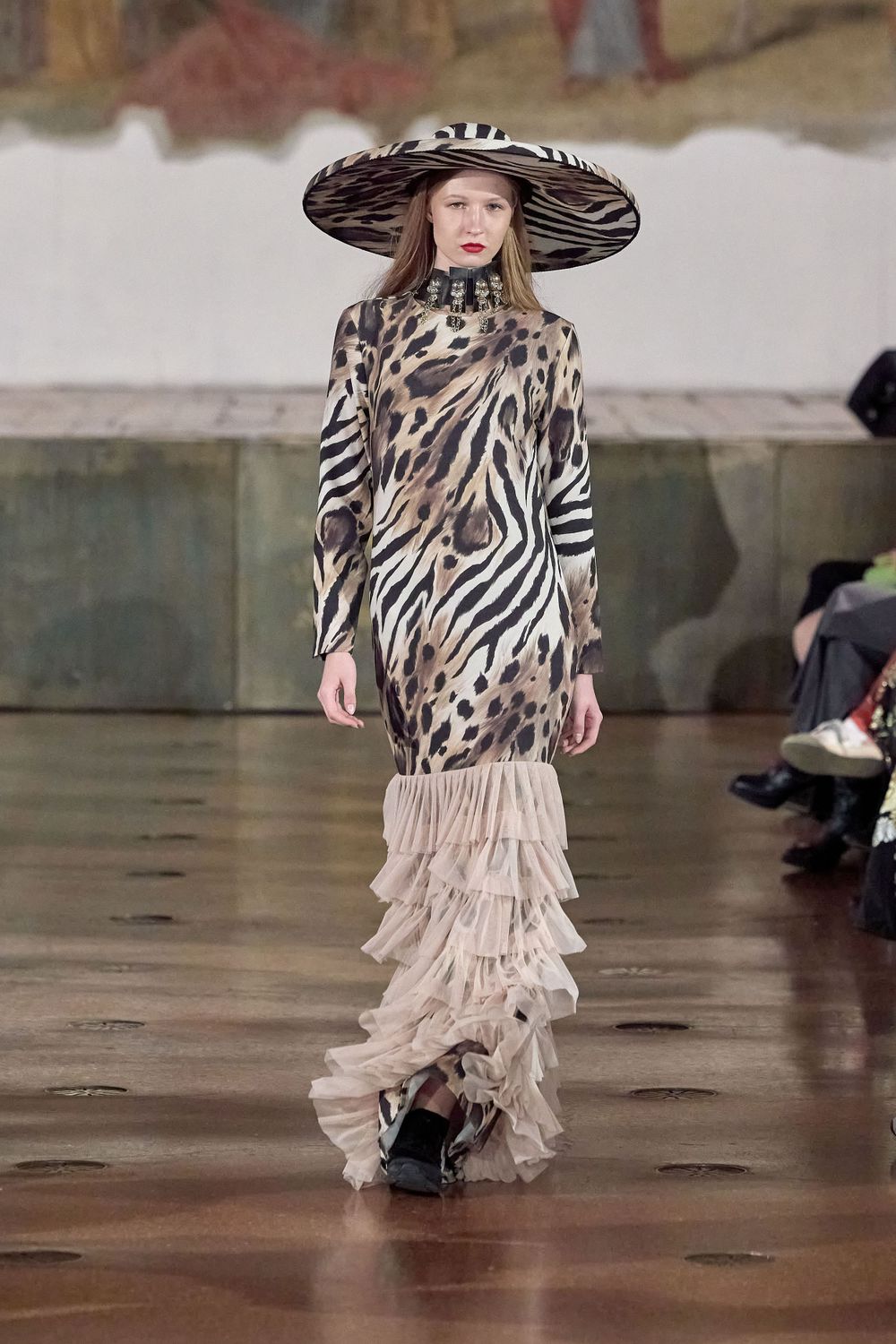 Runway model in zebra print dress with dramatic wide brim hat and ruffled hem.