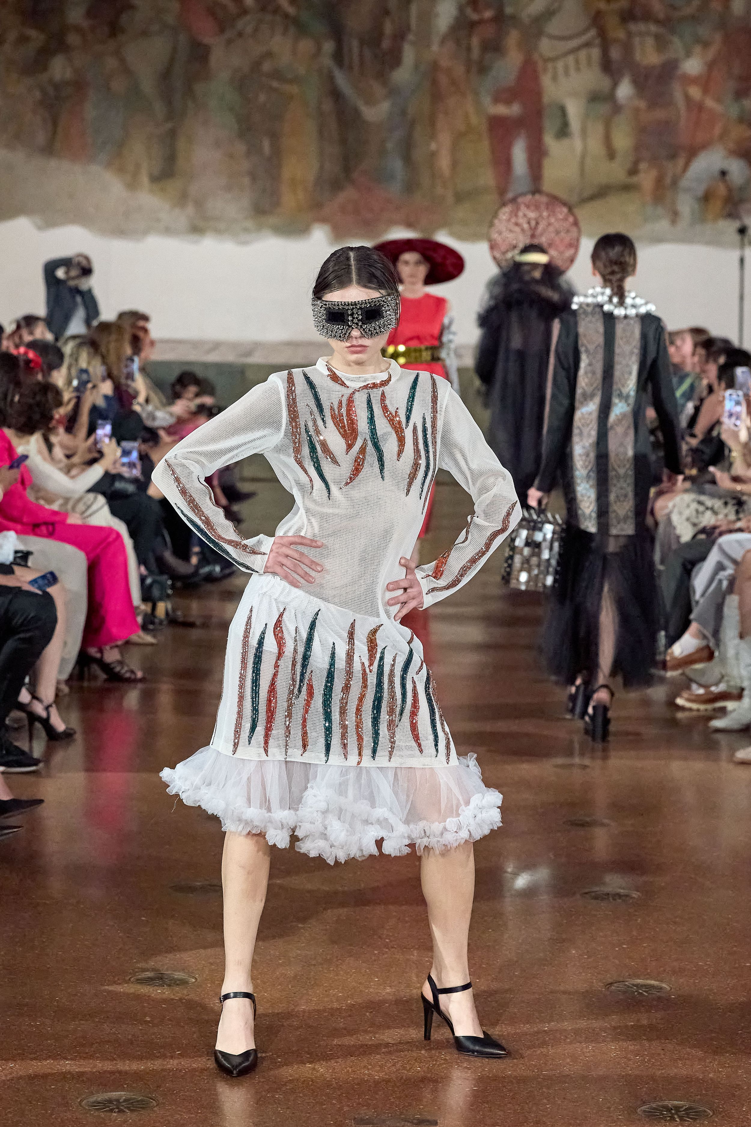 Fashion model presents white dress with colorful stripes and ruffled hem on runway during fashion show.