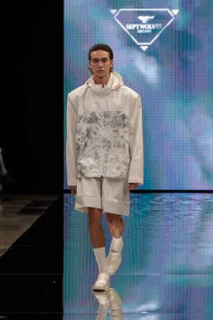 Model presents all-white ensemble with graphic hoodie and shorts on reflective runway.