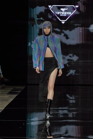 Model walks runway in iridescent jacket and asymmetrical black skirt with boots.