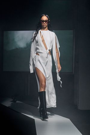 Flowing white asymmetrical dress with dramatic cutouts styled with knee-high boots on runway.