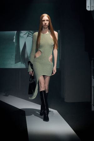 Olive green knit dress with cutouts paired with tall black boots on dimly lit catwalk.