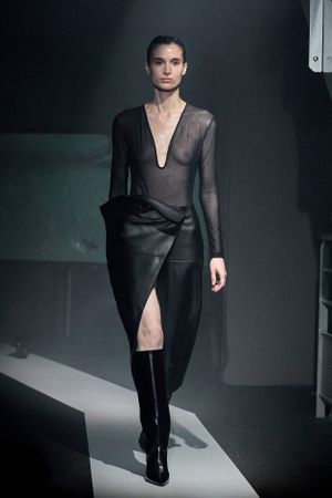 Dramatic black asymmetrical dress with sheer mesh top shown on illuminated fashion runway.