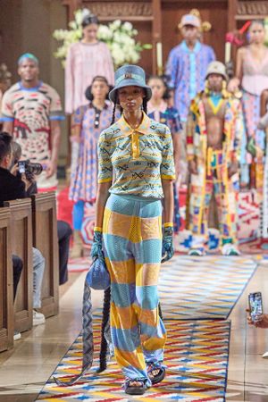 Runway model wearing striped yellow and blue outfit with bucket hat on patterned catwalk.
