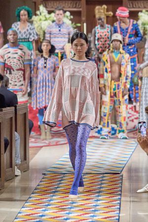 Model showcases oversized pink sweater and patterned tights on colorful geometric runway.