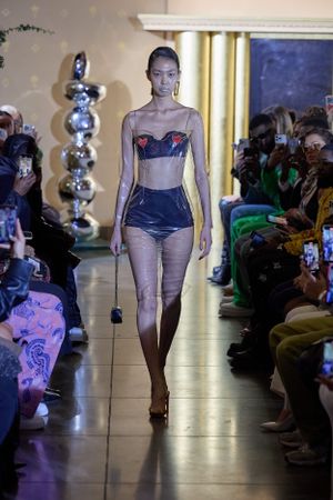 Model showcases blue corset top with shorts on illuminated catwalk with audience in background.