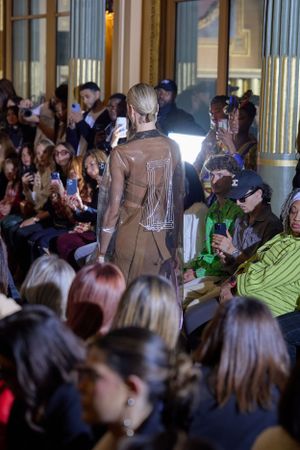 Brown designer outfit with metallic details shown from behind on crowded runway.