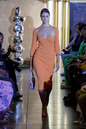 Sleek orange fitted dress presented on indoor runway with seated audience.