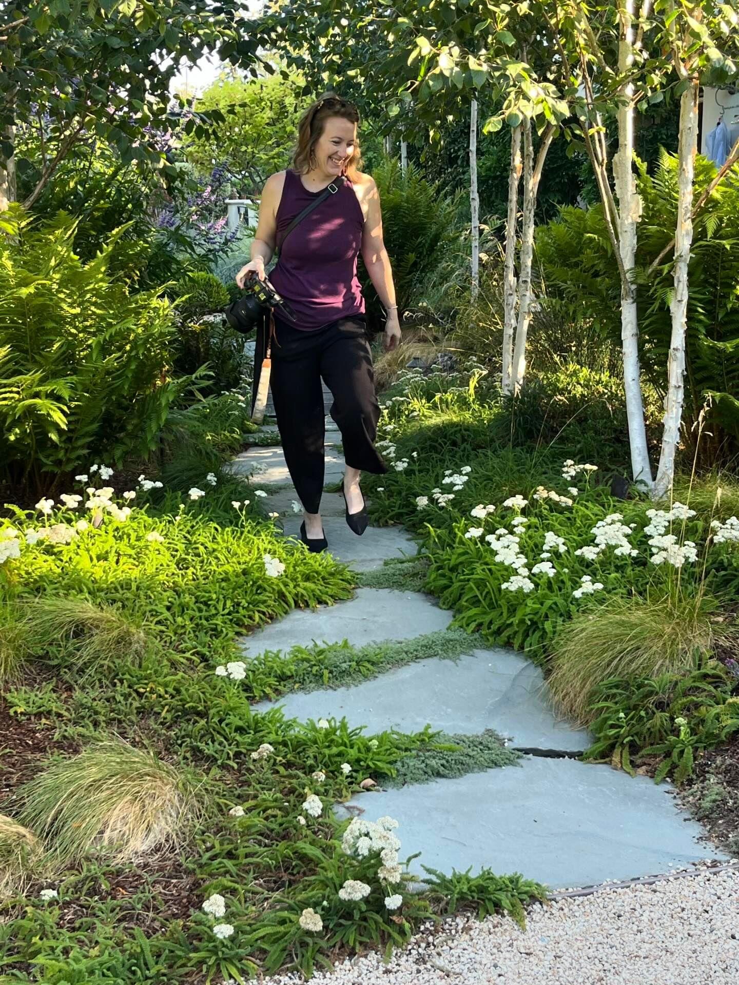 Person walks along curved stepping stones through a lush garden with white flowers and birch trees.