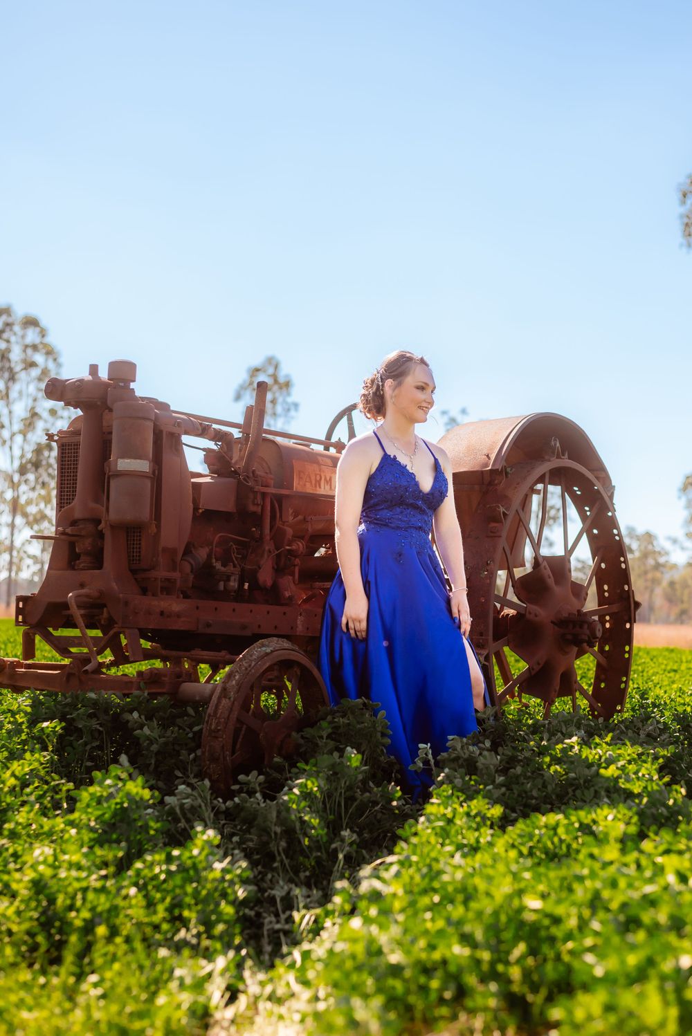 A stunning blue gown contrasts with an antique farm tractor in a sunlit field.