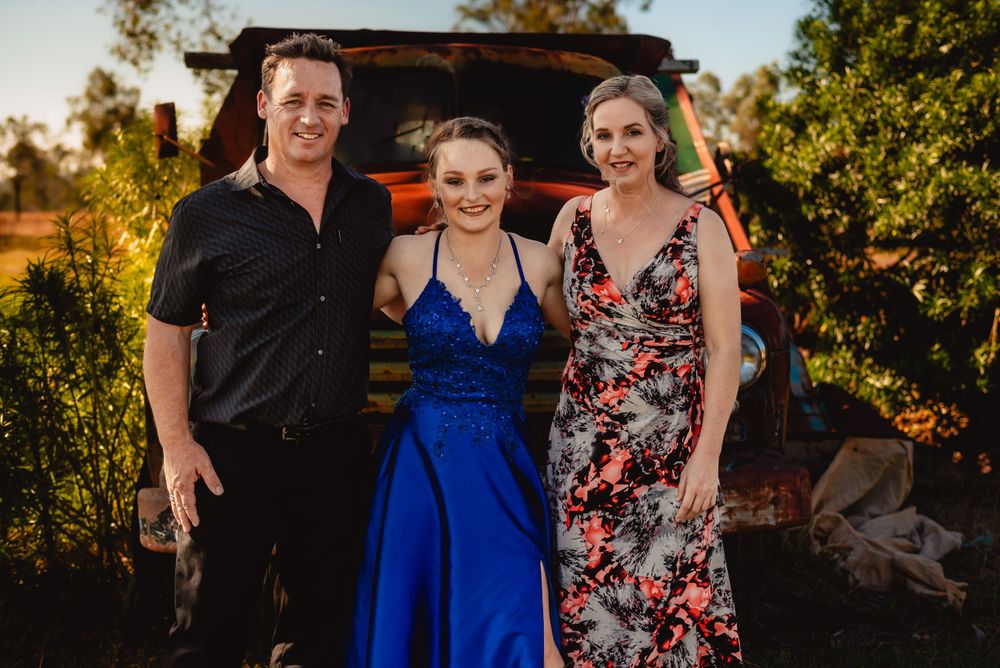 Three people in formal attire pose together in front of rustic truck at sunset.