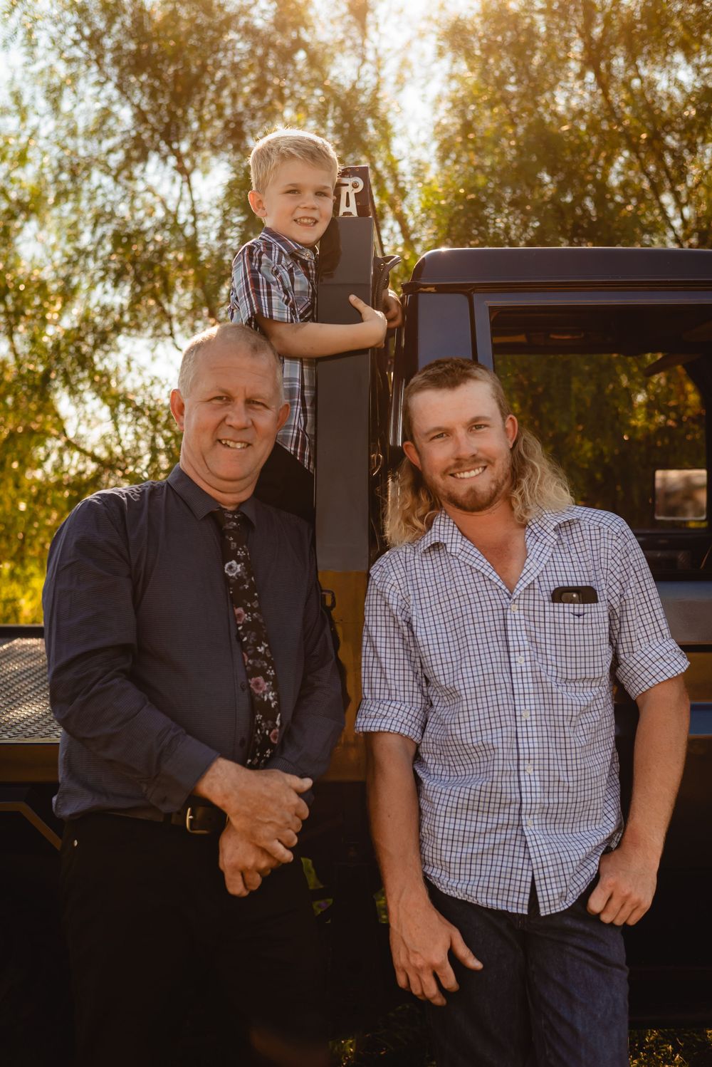 Three generations pose together on vintage truck during golden hour sunset.