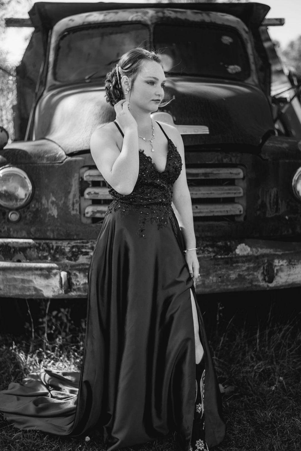 Person in elegant black dress poses dramatically against vintage truck in black and white photo.