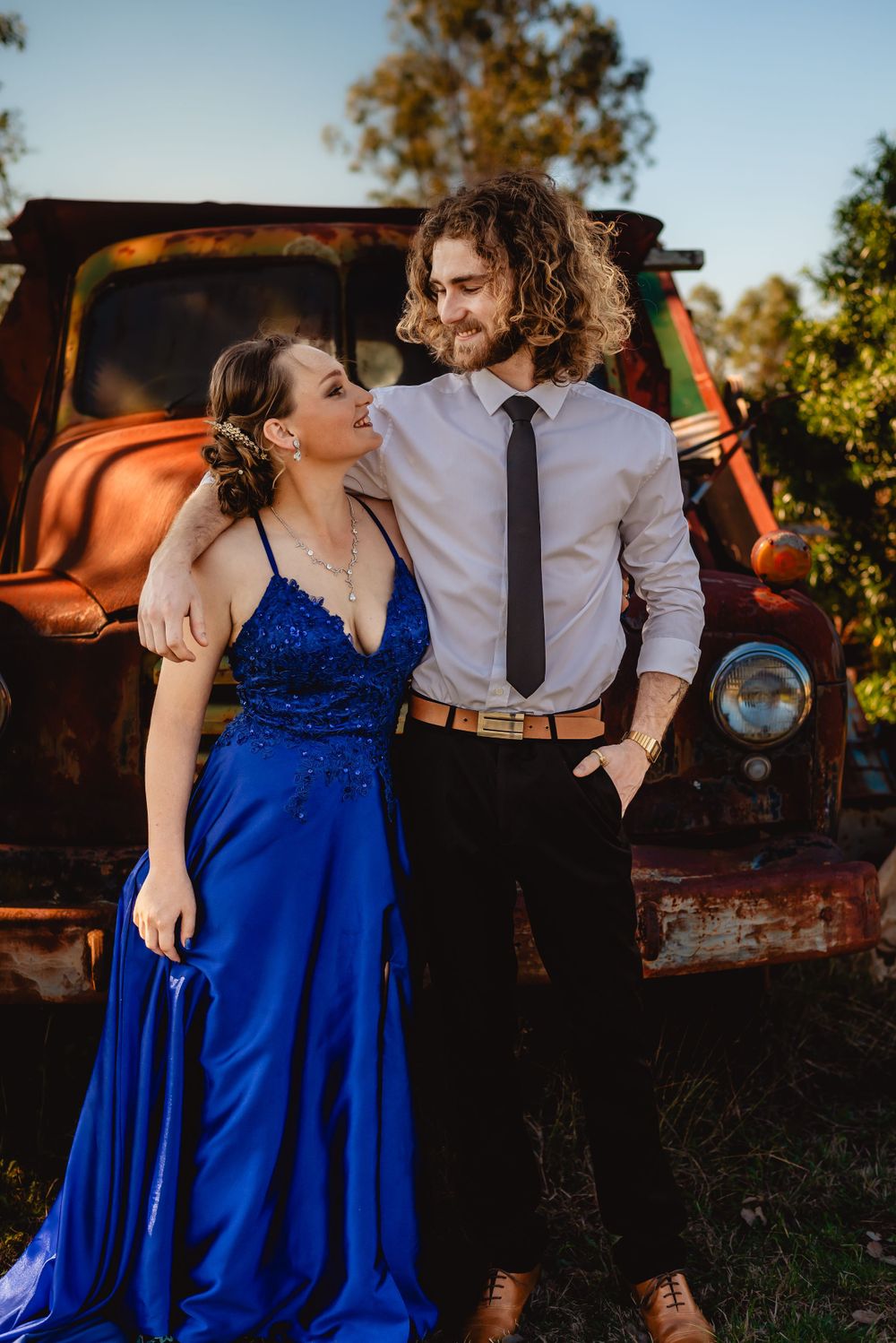 Romantic couple pose by vintage truck, contrasting formal blue gown with business casual attire.