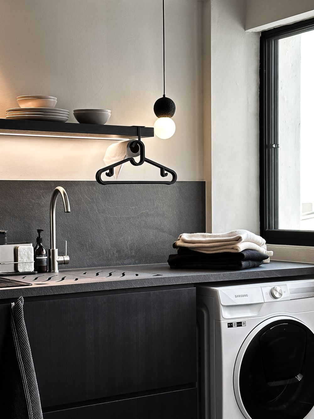 Modern laundry area with black countertops, pendant light, and white washing machine below floating shelves.