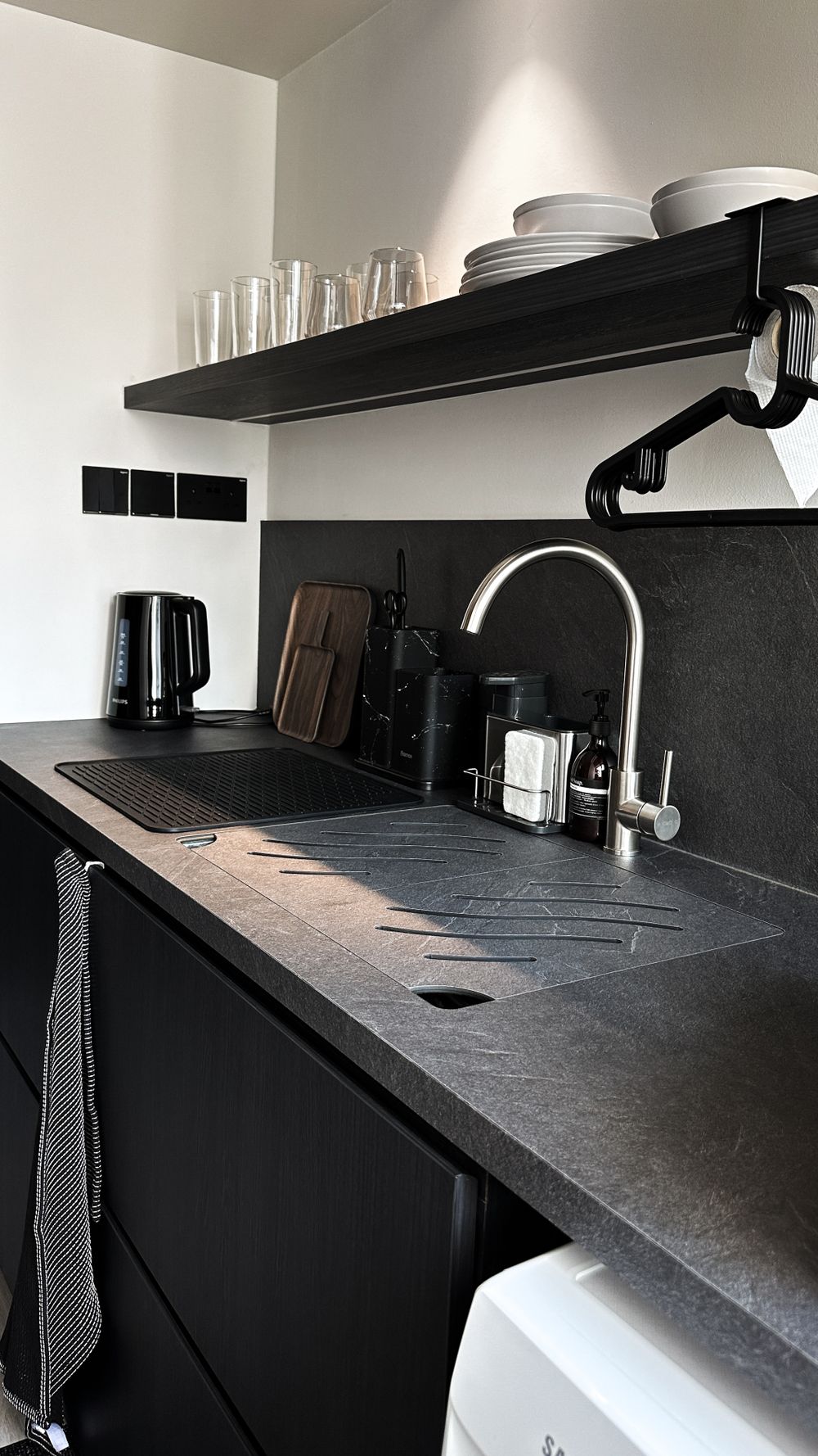 Modern black kitchen with slate countertops, floating shelves, and white dishware display.
