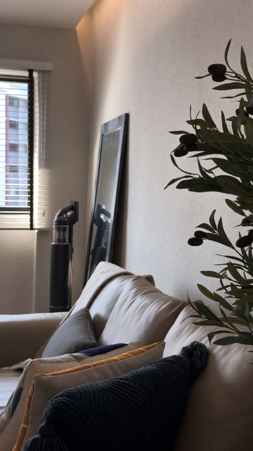 Living room corner with beige couch, window blinds, and decorative plant silhouette.