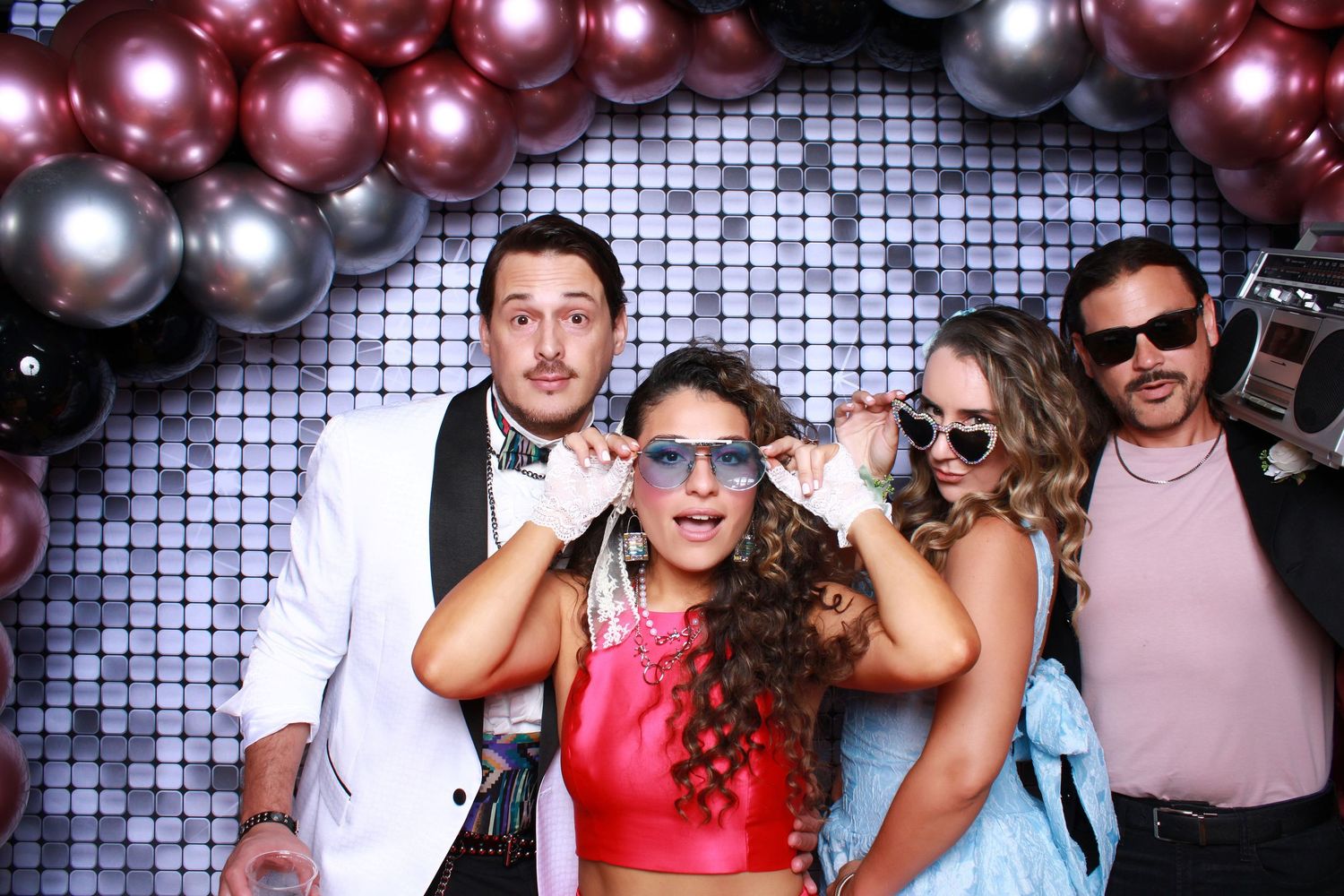 Party scene with three people posing at a photo booth with fun props against a sparkly silver sequin backdrop.