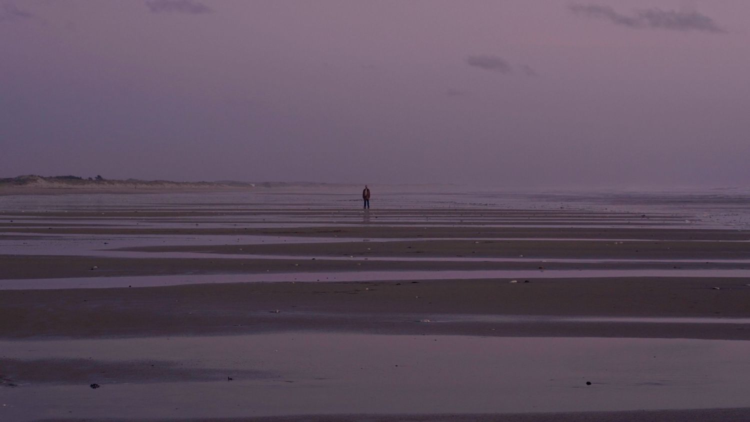Une figure solitaire se dresse sur une vaste plage vide au crépuscule avec un ciel violet spectaculaire.