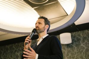Speaker presenting at a business event under circular ceiling lights.