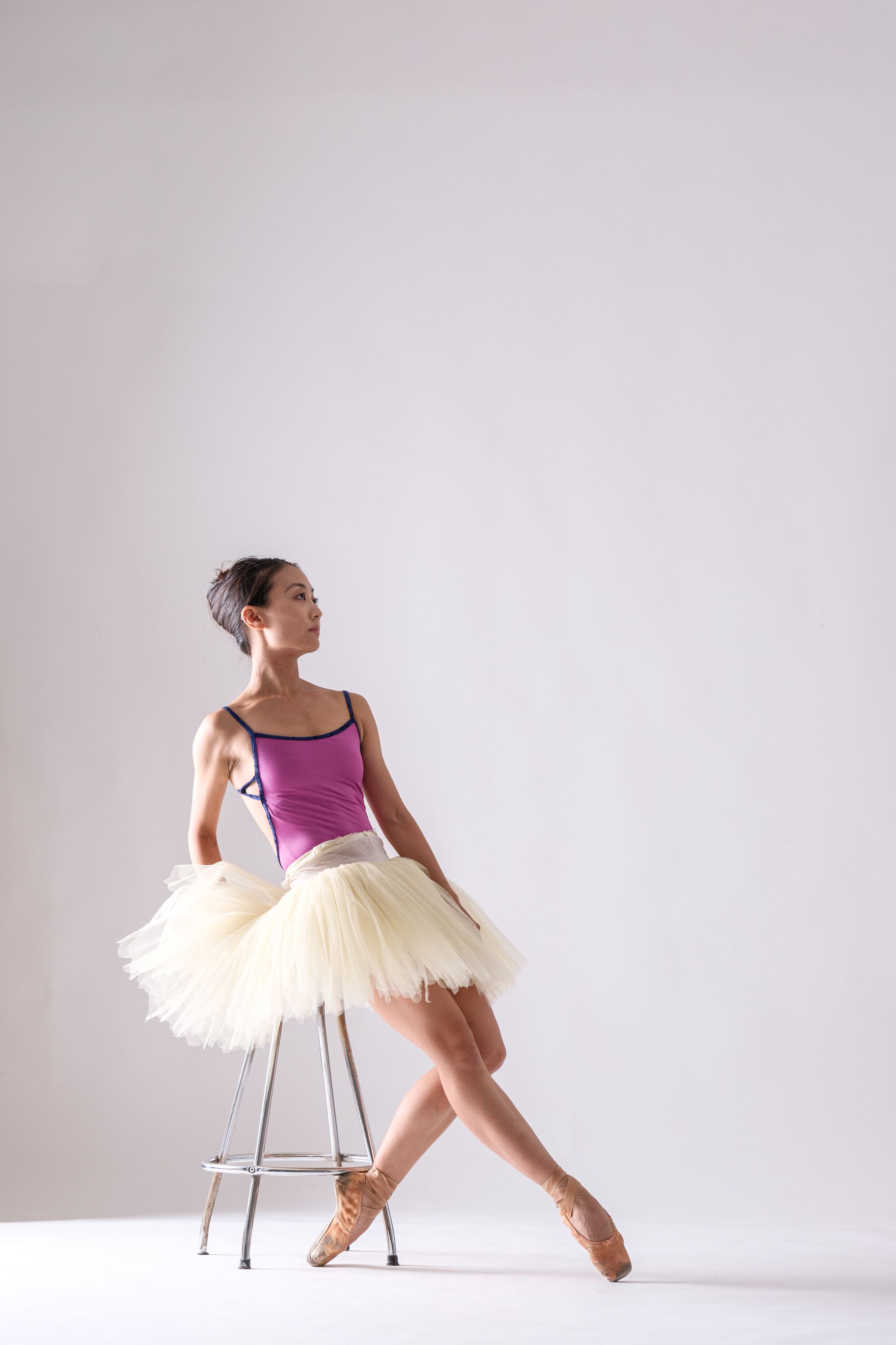 Ballerina in purple top and cream tutu performs elegant movements with stool prop.