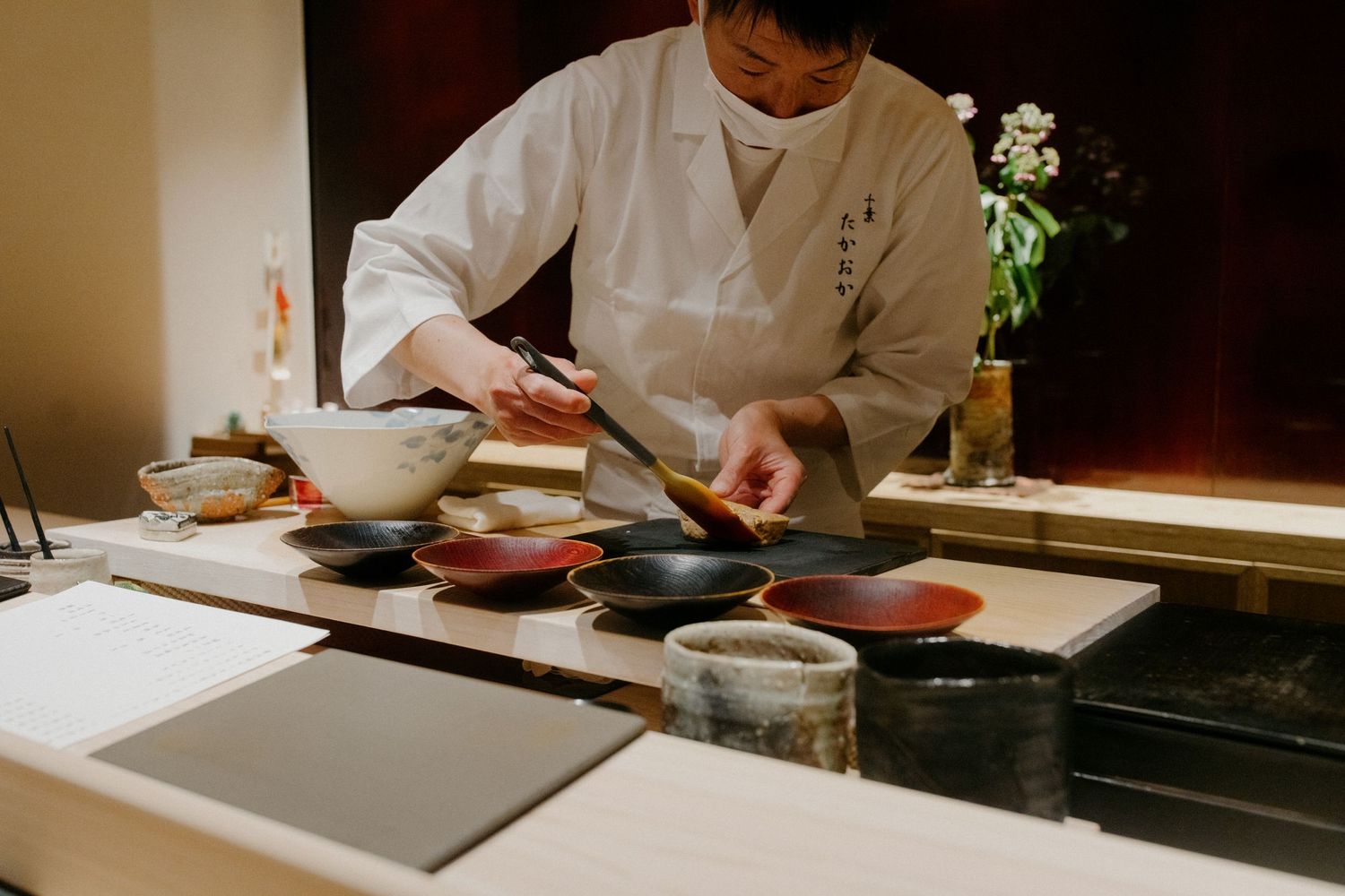 A chef in white uniform prepares dishes at a traditional Japanese counter with small plates and bowls.