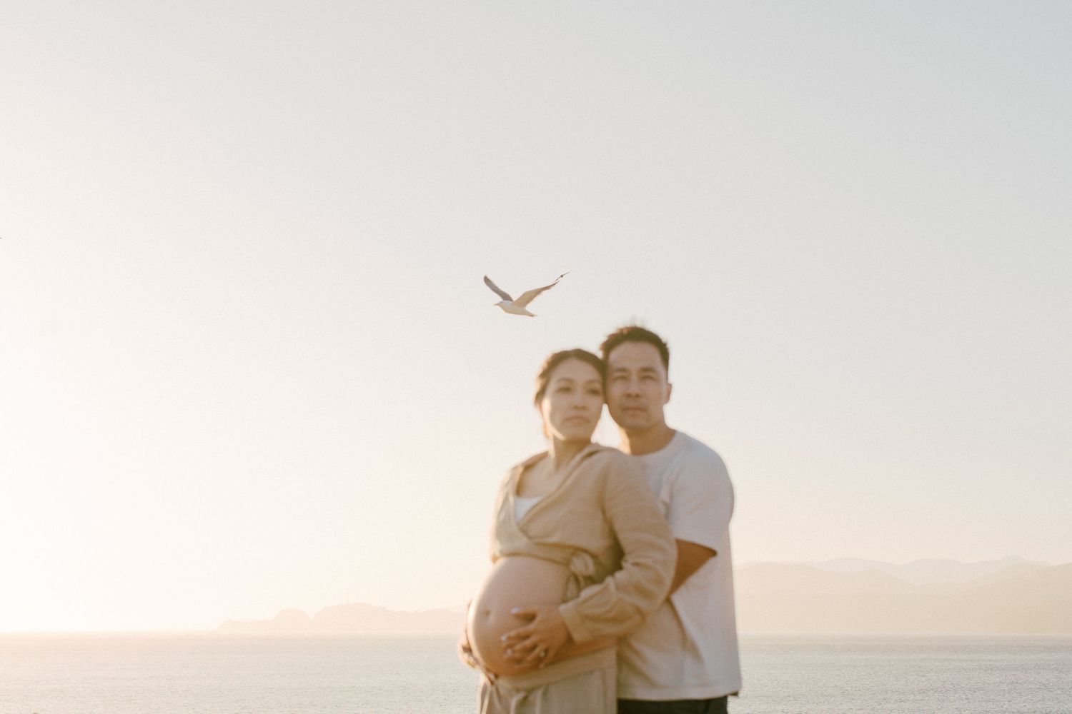 A couple sharing an intimate moment at the beach during sunset with a seabird flying overhead.