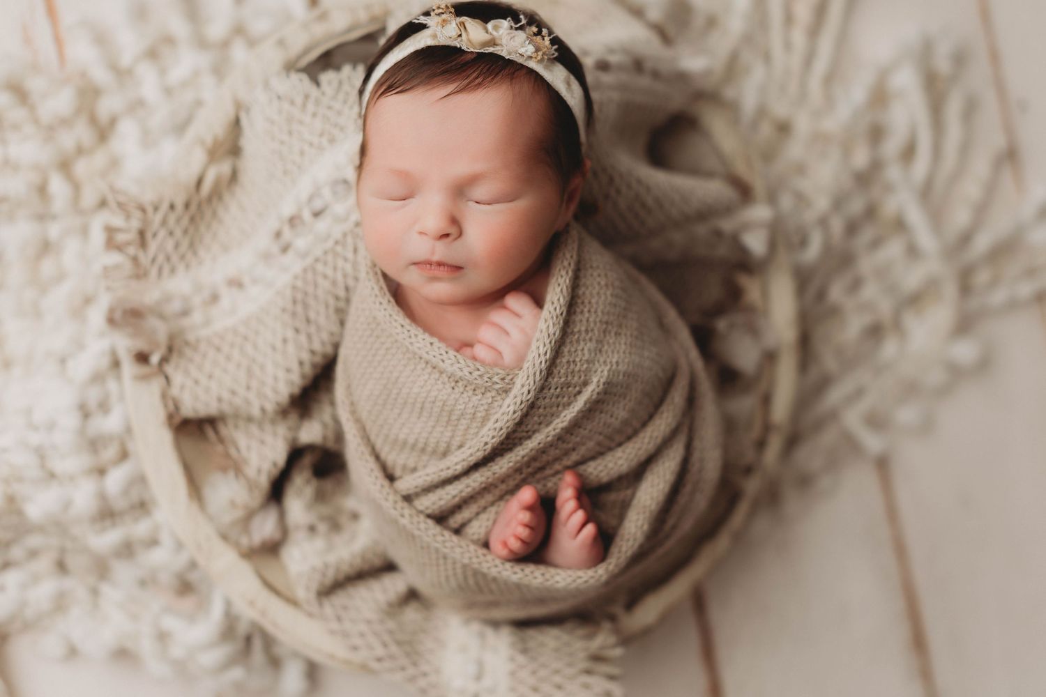 Sleeping newborn wrapped in tan swaddle blanket wearing delicate floral crown on white background.