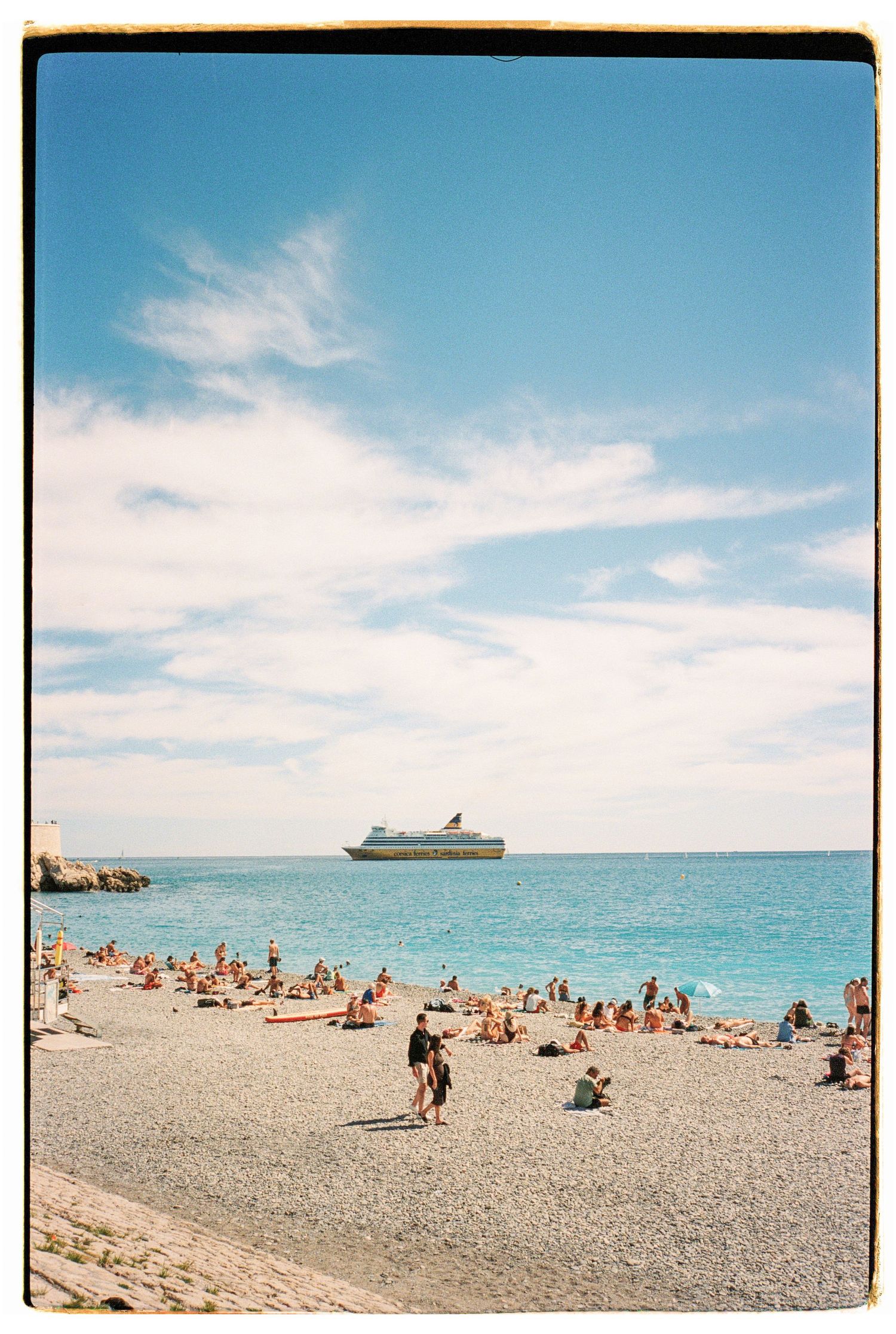 A cruise ship floats on the horizon as beachgoers enjoy a pebbled Mediterranean shore.